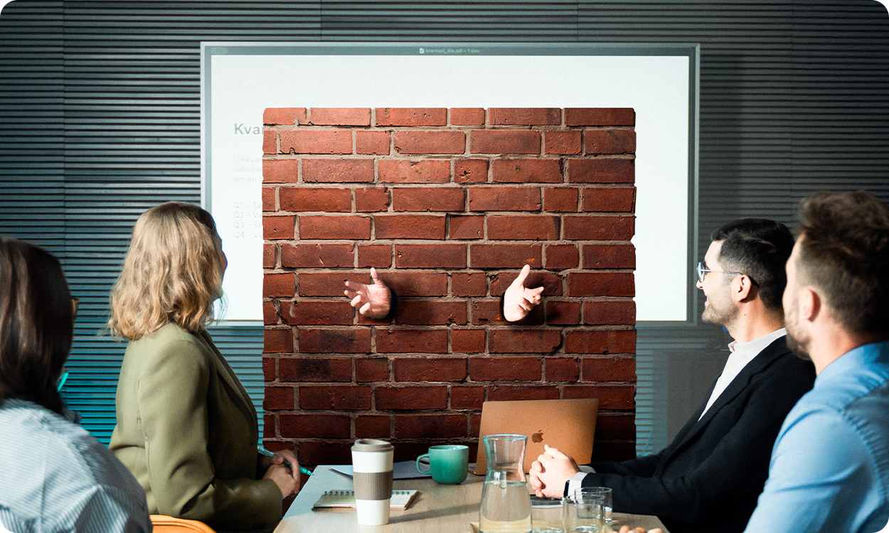 Palaveritilanne viiden ihmisen kesken. Yksi ihmisistä on tiilistä tehdyn "kielimuurin" sisällä.